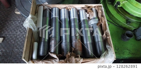 A crate of newly manufactured flange studs, steel threaded rods, with industrial machinery in the background. Close-up of metal flange studs, packaged in a wooden box for transport. Metal studs A crate of newly manufactured flange studs, steel threaded rods, with industrial machinery in the background. Close-up of metal flange studs, packaged in a wooden box for transport. Metal studs 128262891