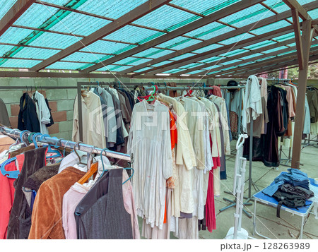 A diverse array of vintage clothing hangs on metal racks under a teal canopy. Garments include dresses, coats, and shirts in various colors and textures. A diverse array of vintage clothing hangs on metal racks under a teal canopy. Garments include dresses, coats, and shirts in various colors and textures. 128263989