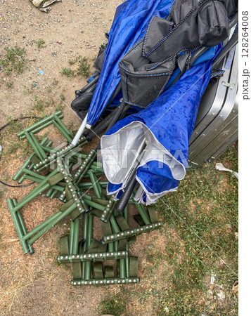 A chaotic arrangement of blue and black umbrellas lies atop green metal stands on a gravel surface, with scattered grass and droplets of water visible. A chaotic arrangement of blue and black umbrellas lies atop green metal stands on a gravel surface, with scattered grass and droplets of water visible. 128264008
