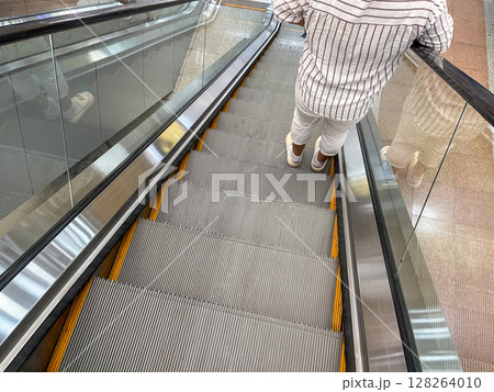 A top-down view of a person descending an escalator. The metallic steps are lined with yellow safety strips, and the individual wears light-colored pants and shoes. 128264010