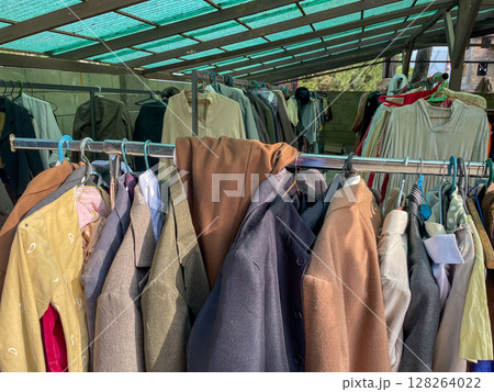 A variety of jackets and coats hang on a metal rack, showcasing colors like beige, brown, and pastel shades under a green-tinted canopy. A variety of jackets and coats hang on a metal rack, showcasing colors like beige, brown, and pastel shades under a green-tinted canopy. 128264022