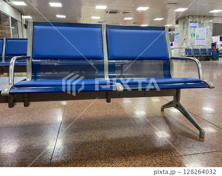 Two blue metal-framed airport seats are positioned in a spacious terminal. The floor features a glossy, speckled pattern, while the background shows empty waiting areas. Two blue metal-framed airport seats are positioned in a spacious terminal. The floor features a glossy, speckled pattern, while the background shows empty waiting areas. 128264032