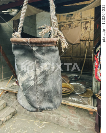 A weathered leather bag hangs from a rope, showcasing scuff marks and a worn texture. Behind it, a rustic wooden door and various metal dishes are visible. 128264033