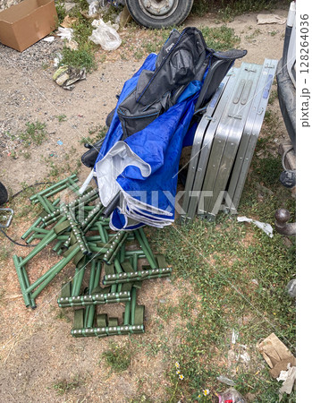 A pile of green metal stands lies on the ground beside a blue tarp and two metallic cases. The area is littered with debris and grass peeking through. A pile of green metal stands lies on the ground beside a blue tarp and two metallic cases. The area is littered with debris and grass peeking through. 128264036