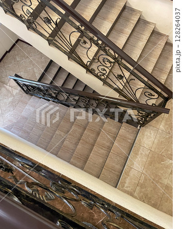 A top-down view of a staircase featuring intricate wrought iron railings. The steps are light-colored tiles, contrasting with the dark railing and wooden accents. 128264037