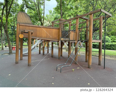 Large wooden play structure featuring a slide and climbing frame resting on a soft rubberized surface in a park setting, surrounded by trees and greenery for children's playtime Large wooden play structure featuring a slide and climbing frame resting on a soft rubberized surface in a park setting, surrounded by trees and greenery for children's playtime 128264324