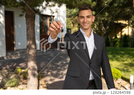Real estate agent presenting house keys and documents on clipboard in front of property for sale 128264598