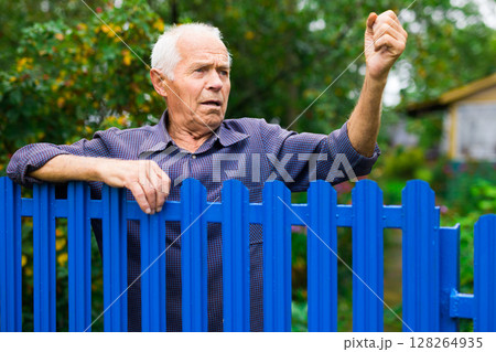 Old man showing fist outdoors beside fence Old man showing fist outdoors beside fence 128264935