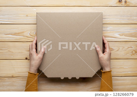 Pizza closed carton box in human caucasian hands on natural wooden table flat lay mockup with blank space 128266079