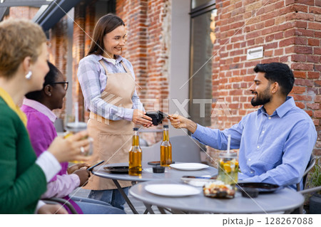 A female waiter is at a table and is asking a customer to pay the bill using a portable payment terminal. Other customers are present. A female waiter is at a table and is asking a customer to pay the bill using a portable payment terminal. Other customers are present. 128267088