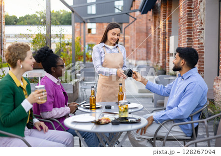 A female waiter assists customers with a portable terminal for payment at an outdoor cafe. 128267094