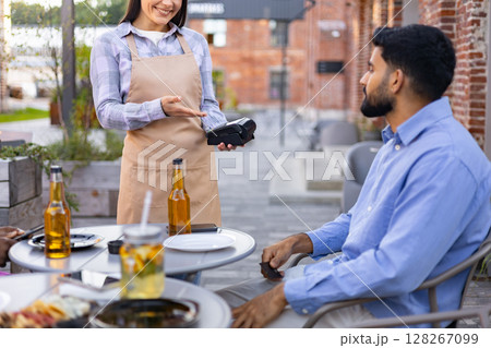 A female waiter offers a payment terminal to a customer at an outdoor restaurant table. A female waiter offers a payment terminal to a customer at an outdoor restaurant table. 128267099