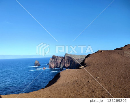 Rugged cliffs rise along a vibrant blue ocean, with waves gently breaking against the rocky formations. Madera, Portugal Rugged cliffs rise along a vibrant blue ocean, with waves gently breaking against the rocky formations. Madera, Portugal 128269295