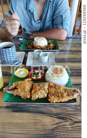 Focus on a plate of fried bangus fish, rice with egg, and sides on green plates with a man eating a plate of rice with stir-fried vegetables. Authentic local Filipino breakfast meal 128269560