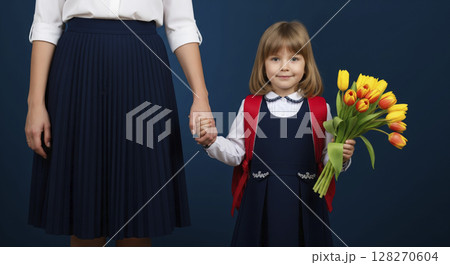 Young schoolgirl in uniform with flowers and backpack holding her mother's hand. First day at school. 128270604