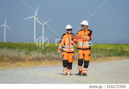 Two wind farm workers in safety orange uniform inspecting wind turbines while walking on gravel path in sunny day 128272560