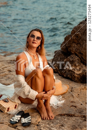 Woman Beach Relaxation: A woman sits on a rocky beach in a white swimsuit and sunglasses. 128273494