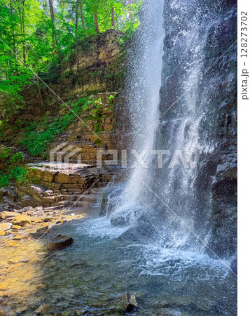 Water flows down rocky cliff creating waterfall in vibrant natural setting. Sunlight filters through trees casting shadows on clear water pool. Concept of eco-tourism, outdoor adventure, relaxation. Water flows down rocky cliff creating waterfall in vibrant natural setting. Sunlight filters through trees casting shadows on clear water pool. Concept of eco-tourism, outdoor adventure, relaxation. 128273702