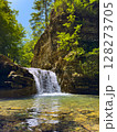 Waterfall pours into tranquil pool, sunlight filtering through leaves. Scenic location in vibrant forest setting with clear water inviting relaxation. 128273705