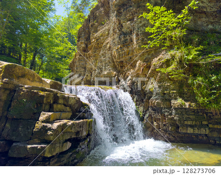 Waterfall flows over rugged rocks in dense forest filled with vibrant greenery. Sunlight shines through tree branches, creating a tranquil ambiance. Concept of nature, outdoor adventure, eco-tourism. Waterfall flows over rugged rocks in dense forest filled with vibrant greenery. Sunlight shines through tree branches, creating a tranquil ambiance. Concept of nature, outdoor adventure, eco-tourism. 128273706