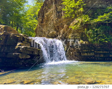 Waterfall pours over rocky ledge creating a tranquil pool below. Sunlight filters through trees, enhancing peaceful atmosphere. Concept of nature escape, outdoor adventures, wellness. Waterfall pours over rocky ledge creating a tranquil pool below. Sunlight filters through trees, enhancing peaceful atmosphere. Concept of nature escape, outdoor adventures, wellness. 128273707