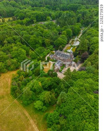 An aerial view of a historic mansion surrounded by lush green trees and a beautifully landscaped garden with a fountain, walking paths, and parking area. The scene shows vibrant summer greenery. An aerial view of a historic mansion surrounded by lush green trees and a beautifully landscaped garden with a fountain, walking paths, and parking area. The scene shows vibrant summer greenery. 128273919