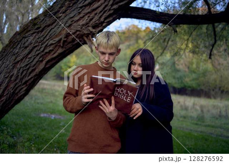 a 15-year-old teenage couple in love reading the holy bible together in the park on a cold autumn evening 128276592