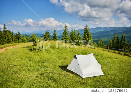 White tourist tent on sunlit, grassy hilltop, surrounded by young pine trees. Panoramic view of lush, rolling mountains under bright blue sky with scattered clouds, creating idyllic camping spot. White tourist tent on sunlit, grassy hilltop, surrounded by young pine trees. Panoramic view of lush, rolling mountains under bright blue sky with scattered clouds, creating idyllic camping spot. 128276621