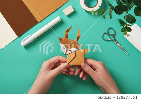 Hands holding a finished paper deer craft. Scissors, tape, glue, and colored paper. Hands holding a finished paper deer craft. Scissors, tape, glue, and colored paper. 128277351