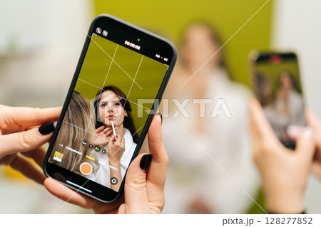 Female hands holding smartphone recording video of beautician applying makeup on model face, creating content for social media or online tutorial in cosmetology clinic. Concept of beauty treatment. Female hands holding smartphone recording video of beautician applying makeup on model face, creating content for social media or online tutorial in cosmetology clinic. Concept of beauty treatment. 128277852