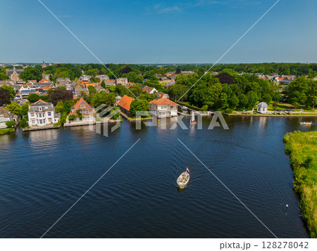 A yacht with a white sail sails along the canal. A beautiful windmill. Aerial view of narrow canal running through lush green fields and a small forest leading to peaceful Dutch village with a marina 128278042