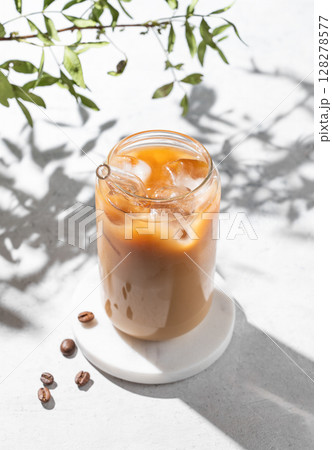 Ice latte coffee in a glass with milk on a light background with coffee beans, branch and shadows. Ice latte coffee in a glass with milk on a light background with coffee beans, branch and shadows. 128278577