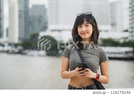 Happy young southeast asian woman teenager blogger using smartphone searching guide. World travel explorer at Chao Phraya River Bangkok asia tourism concept for local cultural experience 128279246