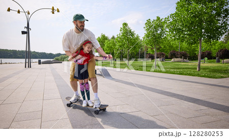 Speed and laughter. Father teaching daughter skateboarding on sunny park pathway Speed and laughter. Father teaching daughter skateboarding on sunny park pathway 128280253