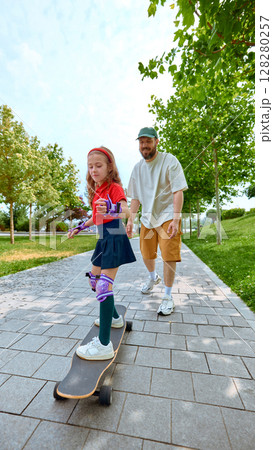 Speed and laughter. Father guiding daughter on skateboard in sunny park 128280257