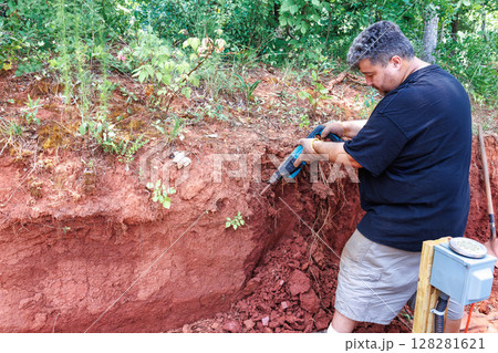 Person uses jackhammer tool to dig into red soil, revealing layer patterns roots in yard full Person uses jackhammer tool to dig into red soil, revealing layer patterns roots in yard full 128281621