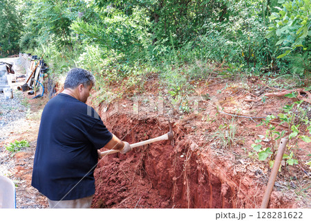 Man is digging red soil near gravel path, surrounded by trees vegetation on work day Man is digging red soil near gravel path, surrounded by trees vegetation on work day 128281622