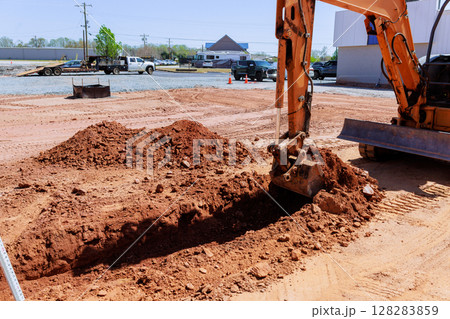 Excavator works on construction site creating trench while trucks are parked nearby during works area 128283859