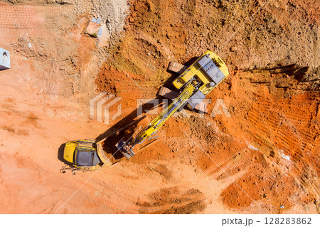 Heavy machinery diging to ground at construction site with reddish dirt surrounding them. Heavy machinery diging to ground at construction site with reddish dirt surrounding them. 128283862