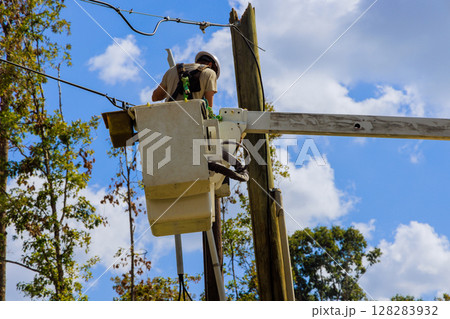 Worker in bucket truck is repairing electrical lines amid trees while sun shines brightly above. 128283932