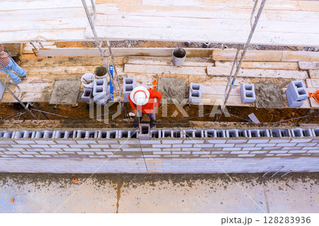 Laborers work on constructing brick wall using scaffolding, blocks, mortar in an active construction area. 128283936