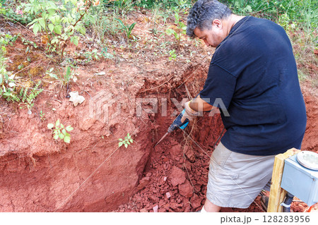 Individual works diligently in rich red soil, using jackhammer tool to break up ground 128283956