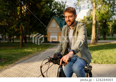 Young man enjoys a sunny day biking through a park with green trees and a residential building nearby Young man enjoys a sunny day biking through a park with green trees and a residential building nearby 128284003