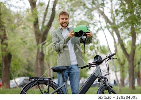Young man smiles while holding a green helmet beside his electric bike in a sunny park Young man smiles while holding a green helmet beside his electric bike in a sunny park 128284005