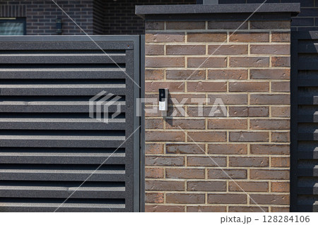 A contemporary intercom is mounted on a brick wall adjacent to a slatted gate in a well-maintained suburban area A contemporary intercom is mounted on a brick wall adjacent to a slatted gate in a well-maintained suburban area 128284106