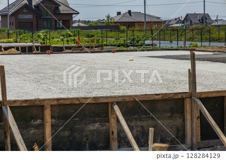 A concrete foundation has been freshly poured at a construction site surrounded by houses and greenery under a bright sky A concrete foundation has been freshly poured at a construction site surrounded by houses and greenery under a bright sky 128284129