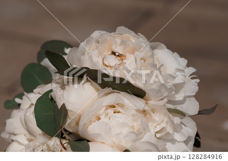 Wedding Rings on White Peonies Bouquet with Eucalyptus, Golden Bands, Romantic Close-Up, Natural Light, Soft Focus Floral Arrangement. Wedding Rings on White Peonies Bouquet with Eucalyptus, Golden Bands, Romantic Close-Up, Natural Light, Soft Focus Floral Arrangement. 128284916