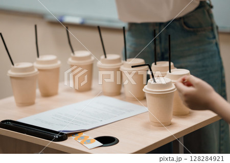 Beverage Sampling Event: Array of Paper Cups and Hand Reaching, Light Wood Table, Denim Jeans, Blank Paper, Neutral Palette, Testing Scenario. Beverage Sampling Event: Array of Paper Cups and Hand Reaching, Light Wood Table, Denim Jeans, Blank Paper, Neutral Palette, Testing Scenario. 128284921