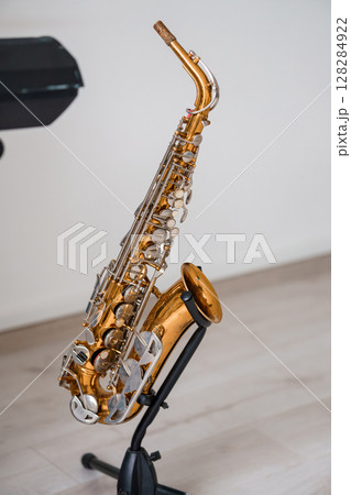 Golden Alto Saxophone on Stand, Music Instrument, Studio Shot, Warm Lighting, Brass Texture, Jazz Performance, Solo Practice, Elegant Design. Golden Alto Saxophone on Stand, Music Instrument, Studio Shot, Warm Lighting, Brass Texture, Jazz Performance, Solo Practice, Elegant Design. 128284922