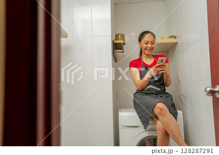 Young Woman Relaxing on Washer, Using Smartphone in Modern Bathroom Setup Young Woman Relaxing on Washer, Using Smartphone in Modern Bathroom Setup 128287291